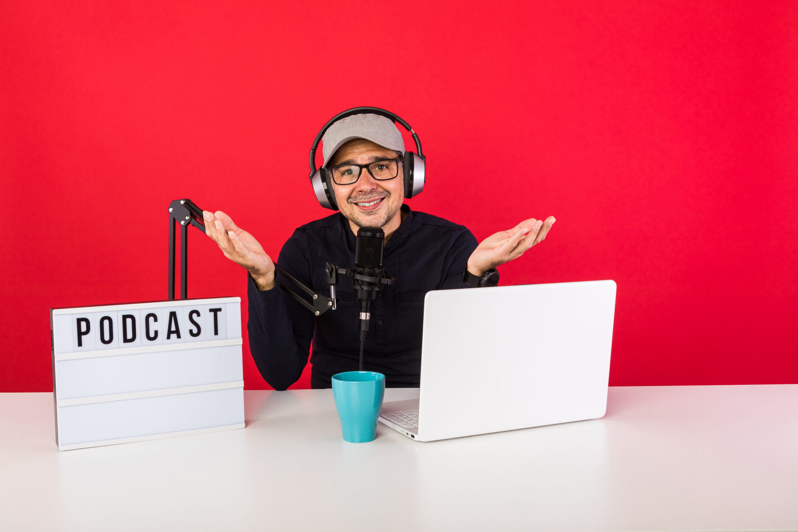 Man presenter with cap in podcast radio recording studio making hand gestures, next to a computer, a microphone and a light box with the word podcast, on red background. Podcasting, broadcast concept Man presenter with cap in podcast radio recording studio making hand gestures, next to a computer, a microphone and a light box with the word podcast, on red background. Podcasting, broadcast concept